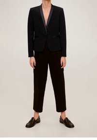Blazer nero con rever in satin, abbinato a pantaloni neri plissettati e mocassini. Il blazer ha un design aderente e non è abbottonato.