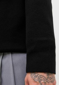 Osoba v černém texturovaném topu s dlouhým rukávem, jejíž tetovaná ruka je viditelná, kombinována se světle šedými plisovanými kalhotami.