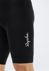 Κοντινό πλάνο μαύρου ποδηλατικού σορτς με λευκό λογότυπο "Rapha" στην εξωτερική πλευρά του μηρού, φορεμένο στο πόδι ενός ατόμου πάνω σε απλό φόντο.