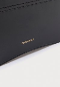 Zwarte leren handtas met een gladde textuur. Heeft een minimalistisch ontwerp en een gouden logo "Coccinelle." Gestikte details langs de rand.