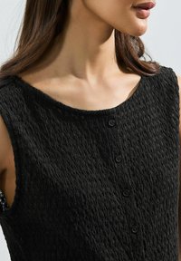 Black sleeveless dress with a textured surface, wide neckline, and five flat buttons in front. Fabric appears lightweight and wrinkled.