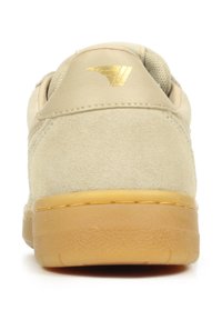 Zapatilla de ante beige con un acabado texturizado, que cuenta con una suela de goma marrón y un acento dorado en el talón. Forma plana y redondeada.