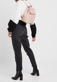 Vaaleanpunainen reppu henkilöllä, jolla on mustavalkoinen neulepaita ja mustat kapenevat housut, yhdistetty mustiin korollisiin nilkkureihin.