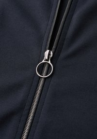 Cremallera con tirador de anilla metálica sobre tela oscura, parcialmente cerrada, con dientes visibles y textura textil suave.