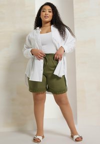 Vit skjorta med knappar över en vit linne, tillsammans med olivgröna shorts med spetsdetaljer i fållen. Modellen bär vita sandaler.