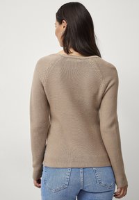 Beige gebreide trui met een ronde hals en ribbelstructuur. Heeft lange mouwen en een licht gebogen zoom, gecombineerd met lichtblauwe jeans.