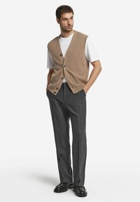 Beige geripptes Weste über einem weißen Hemd, kombiniert mit grauen maßgeschneiderten Hosen und schwarzen Schuhen. Besitzt Knöpfe und einen lockeren Schnitt.