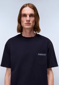 Marineblå kortærmet t-shirt med rund halsudskæring og en påsat lomme. Har et logo på lommen og er lavet af bomuld. Glat tekstur.
