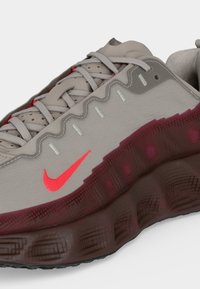 Γκρι και κόκκινο sneaker Nike με κυματιστές ραφές, διαφανή βαθυκόκκινη σόλα και κόκκινο λογότυπο Nike στο πλάι.