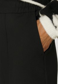 Mustat housut, joissa on sileä kangas ja sivutaskut. Vyötärökin on joustava, mikä tuo mukavuutta. Ylhäällä näkyy valkoinen villapaitatakki.