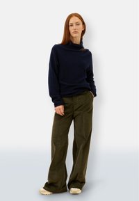 Navyblauer Rollkragenpullover mit lockerer Passform, kombiniert mit olivgrünen weit geschnittenen Cordhosen. Das Model trägt helle Sneakers.