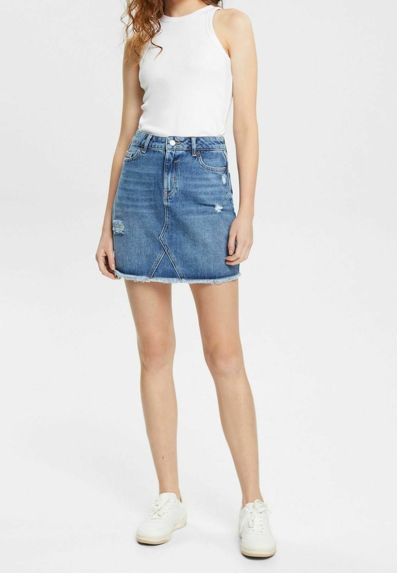 Jupe mini en jean bleu, avec un design déchiré, un ourlet brut et des poches avant. Associée à un haut blanc sans manches et des baskets blanches.