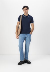 Navy polo shirt with white accents, light blue jeans, and black shoes. Model holds a tan shoulder bag, standing against a white background.