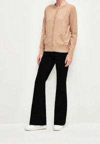 Cardigan beige avec col rond, fermeture à boutons et deux poches avant ; associé à un pantalon noir évasé et des baskets blanches.