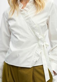 Blouse blanche à volants nouée sur le côté, portée avec un pantalon plissé taille haute vert olive.