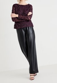 Haut en dentelle bourgogne avec un motif floral texturé, encolure ronde et manches longues ; associé à un pantalon large en simili cuir noir et des sandales noires.