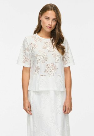 Femme portant un haut en dentelle blanche à manches courtes et une jupe assortie avec des motifs floraux, debout devant un fond clair uni.