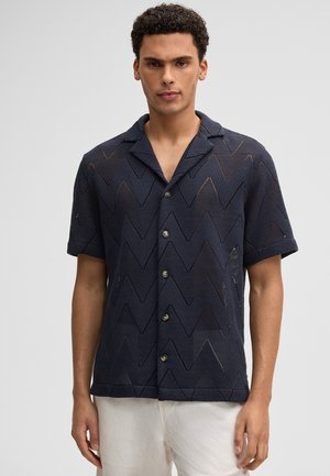 Mężczyzna w granatowej, krótkorękawowej koszuli zapinanej na guziki z wzorem w zygzak oraz jasnych spodniach, stojący na jednolitym tle.