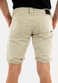 Beige katoenen shorts met opgerolde zomen, met twee achterzakken en een zwart leren patch bij de taille. Glad oppervlak met subtiele stiksels.