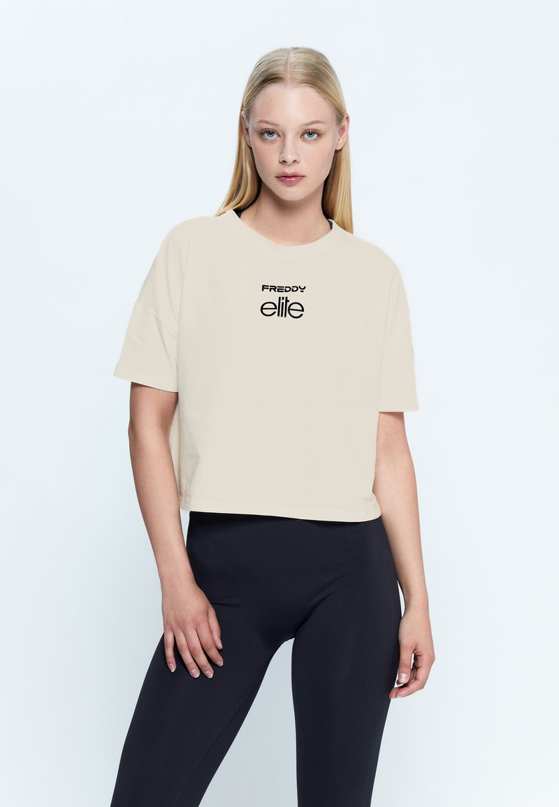 Camiseta recortada color beige de tejido suave con mangas cortas, que presenta un logo negro "FREDDY elite" impreso en el frente.