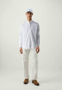 Weißes langärmliges Hemd mit Knöpfen, hellblaues Cap, cremefarbene Hose und braune Stiefel. Einfaches Design, glatte Textur, minimale Markenkennzeichnung auf dem Hemd.