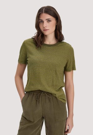 Frau mit schulterlangem braunem Haar, die ein grün-gelb gestreiftes kurzärmeliges Shirt und olivgrüne Hosen mit Kordelzug trägt.