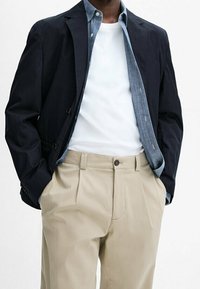 Blazer blu navy sopra una camicia di jeans azzurro chiaro, una t-shirt bianca e pantaloni beige con pieghe. Il blazer presenta due bottoni e una vestibilità personalizzata.