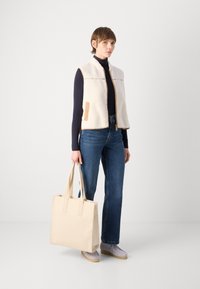 Fleece vest in cream with beige accents, paired with blue jeans and light gray shoes, holding a beige tote bag.