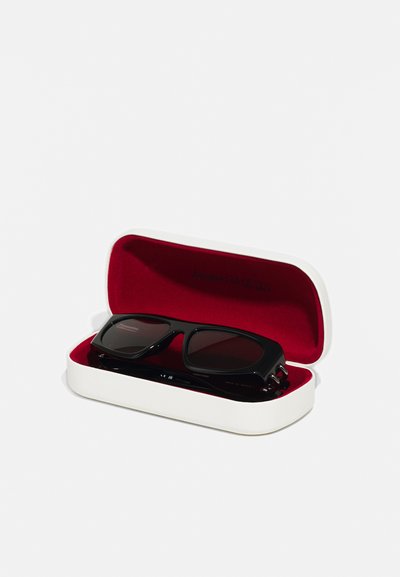 Occhiali da sole neri con lenti scure in un astuccio bianco con interno rosso. L'astuccio è rettangolare con una superficie liscia e il logo del marchio all'interno.