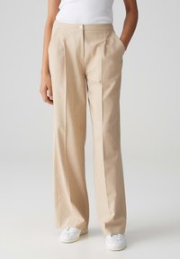Beige wijde broek van lichtgewicht stof, voorzien van een middenplooi, zijzakken en een enkele knoop aan de voorkant. Draagt bij witte sneakers.