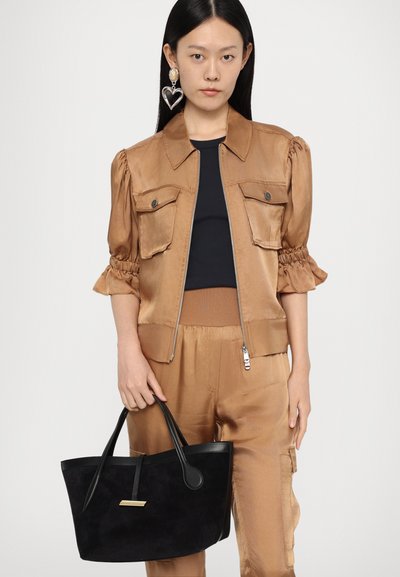 Braune Satinjacke mit Puffärmeln, zwei Fronttaschen, kombiniert mit einer schwarzen Wildledertasche mit goldenen Akzenten. Model hält die Tasche.