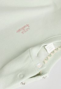 Lysegrøn babytrøje med en blød tekstur, der har pink broderet tekst og en knaplukning ved kraven med et mønstret indvendigt foer.