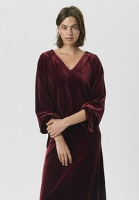 Burgunderfarbenes Samtkleid mit V-Ausschnitt und langen, gerafften Ärmeln. Weicher Stoff, mittelange Gestaltung mit lässiger Passform.