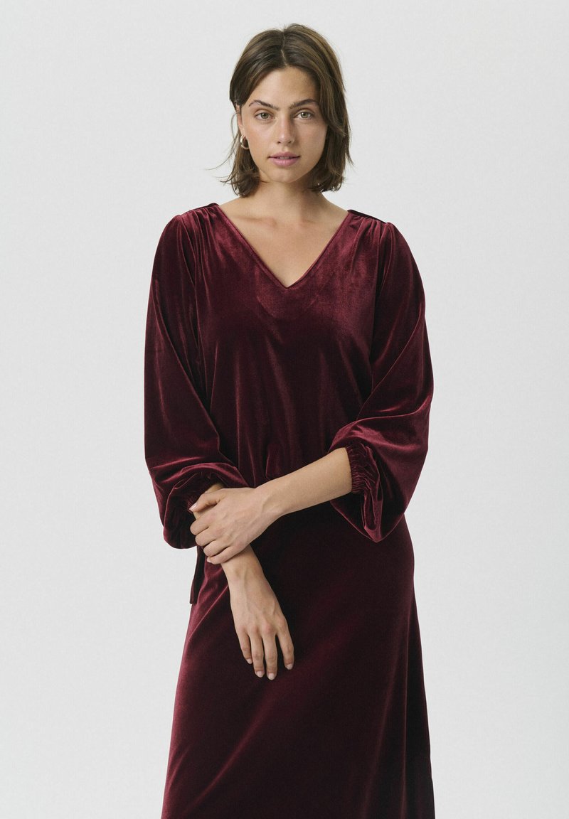 Burgunderfarbenes Samtkleid mit V-Ausschnitt und langen, gerafften Ärmeln. Weicher Stoff, mittelange Gestaltung mit lässiger Passform.