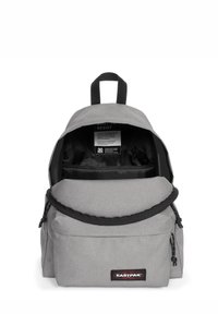 Mochila de tela gris con un bolsillo frontal con cremallera, correas ajustables y un asa reforzada. Forro interior negro y etiquetas visibles en el interior.