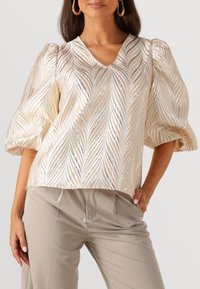 Lichtgouden blouse met een V-hals, voorzien van gewatteerde bladranden en pofmouwen. Gecombineerd met beige broeken voor een complete look.