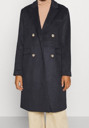 Manteau en laine bleu marine foncé croisé avec quatre boutons, larges revers et poches à rabat, porté sur un col roulé beige et un pantalon clair.