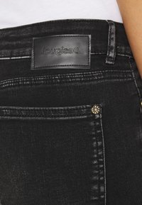 Acercamiento al bolsillo trasero de unos pantalones de mezclilla negros, con una etiqueta de marca de cuero cosida y detalle de remache metálico cerca del borde.
