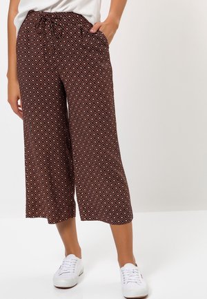 Donna che indossa pantaloni marroni ampi e corti con motivo geometrico a diamante, sneakers bianche e top bianco, in piedi con una mano in tasca.