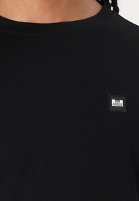 Black cotton t-shirt featuring a round neckline, short sleeves, and a small square label with "WEEKEND OFFENDER" on the chest.