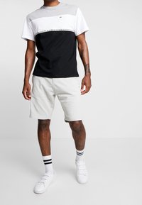 Camiseta con paneles grises, blancos y negros; presenta un detalle del logo. Combinada con pantalones cortos gris claro. Lleva zapatos blancos y calcetines a rayas negras.