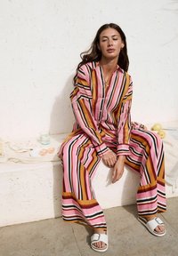 Set di abbigliamento da casa a righe con tonalità di rosa, arancione e nero. Top ampio con maniche lunghe e bottoni, pantaloni a gamba larga. Includendo ciabatte bianche.