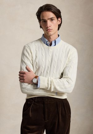 Jeune homme aux cheveux foncés portant un pull en maille torsadée blanc sur une chemise bleue et un pantalon en velours côtelé marron, debout les bras croisés.