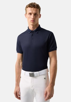 Young man wearing a fitted navy blue polo shirt and white pants with a white belt, standing against a plain background.