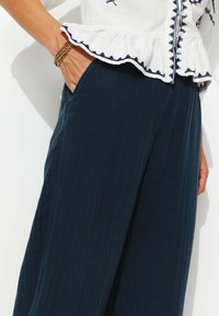 Pantalons larges bleu marine avec une texture douce, dotés de poches latérales et d'une ceinture lisse. Associés à un chemisier blanc avec des accents brodés.