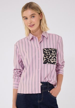 Kvinde iført en pink stribet skjorte med leopardprintlomme, kombineret med mørkeblå jeans, står med den ene hånd i lommen.