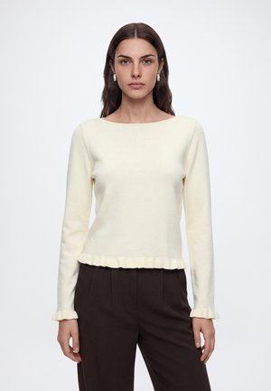 Femme aux longs cheveux foncés portant un haut crème à manches longues avec des volants aux poignets et à l'ourlet, associé à un pantalon brun foncé, sur un fond uni.