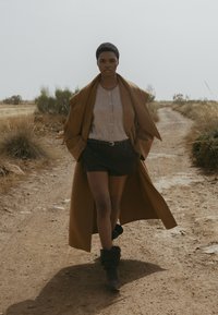 Brauner Wollmantel, kurze schwarze Shorts, graues Hemd und graue Knöchelstiefel; geht auf einem Schotterweg, umgeben von spärlicher Vegetation.