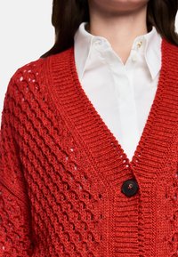 Roter Strickcardigan mit V-Ausschnitt, offenem Strickmuster und großem schwarzem Knopf vorne. Darunter ein weißes Hemd mit Kragen.