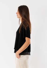 Top nero a maniche corte con design a strati, caratterizzato da un orlo inferiore trasparente. Abbinato a pantaloni beige a gamba larga, con una vestibilità comoda.